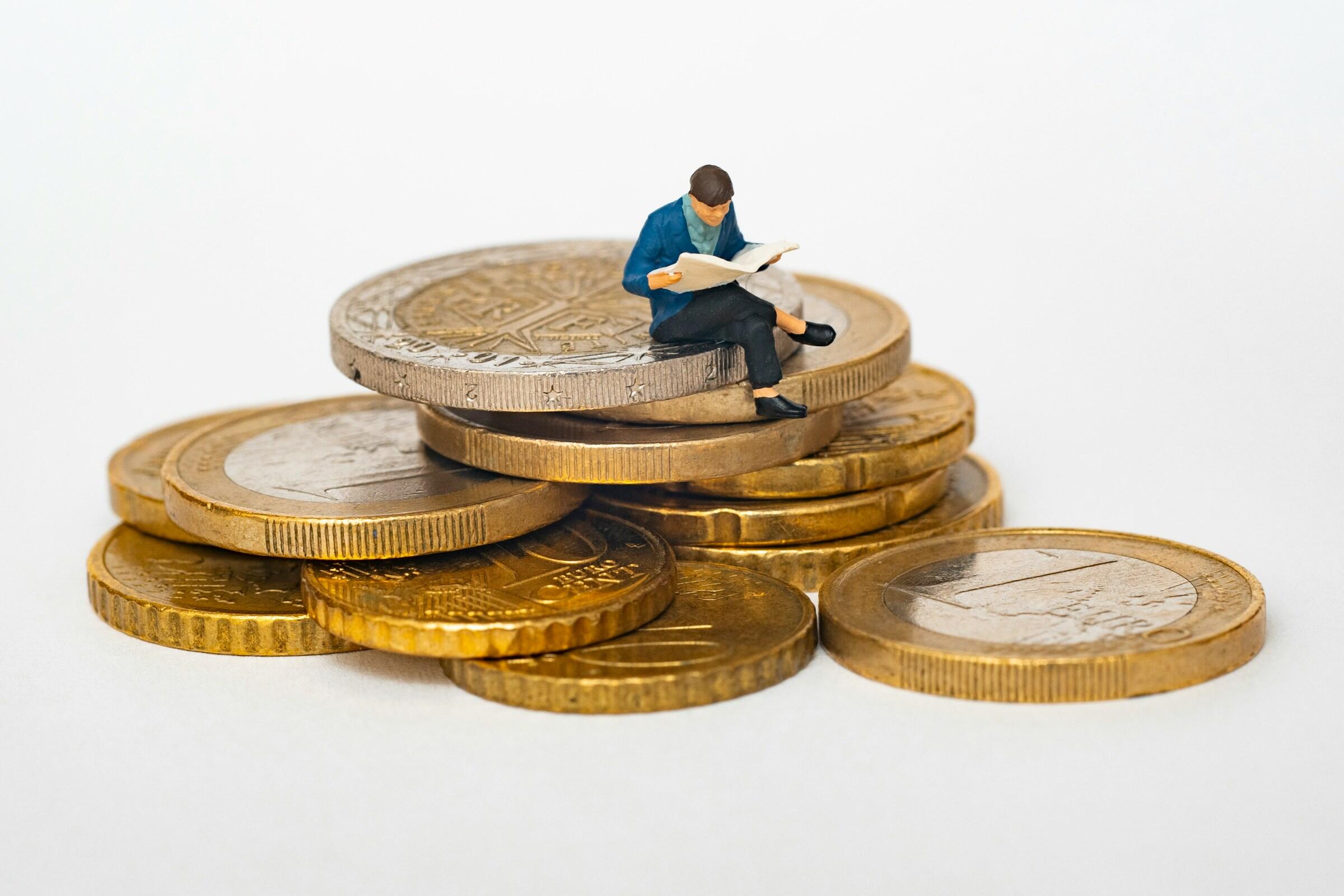 Eine Miniaturfigur mit einem Buch sitzt auf einem Stapel Münzen.